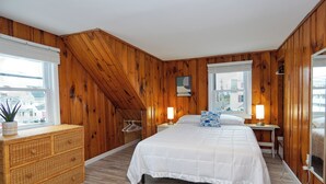 Een slaapkamer met een houten schuine plafond, een groot bed met witte beddengoed, twee bedlampen, een dressoir en een raam met uitzicht op gebouwen.