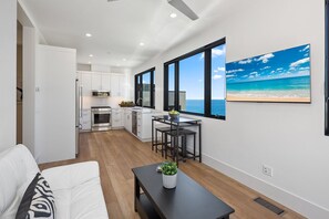 Een moderne woonkamer met een witte bank, een donkere salontafel met een plant, en een keuken met witte kasten en een groot raam met uitzicht op de oceaan.