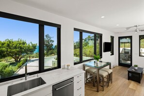 Een moderne keuken met grote ramen, een eethoek met een glazen tafel en uitzicht op groen en een zwembad.