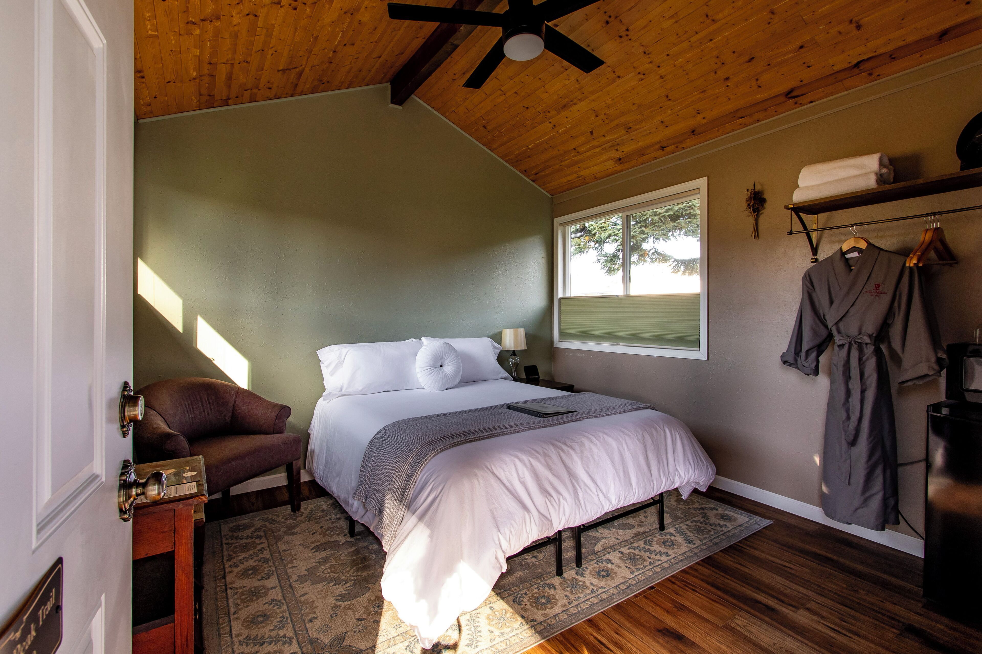 A bedroom with a large bed, a wooden ceiling, a window, a robe hanging on a rack, and a small table with a lamp.