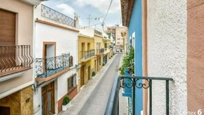Uma rua estreita com edifícios em ambos os lados, com varandas e grades de metal, e plantas em vasos.