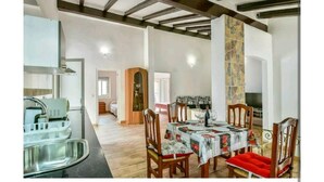 Uma sala de jantar com mesa posta para quatro pessoas, cadeiras de madeira e um banco vermelho com almofadas. Uma área de cozinha com pia e fogão é visível à esquerda.