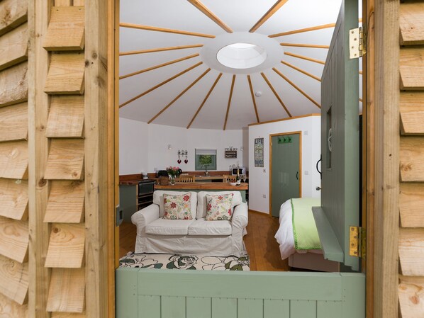 A cozy interior with a wooden ceiling, a white sofa with floral cushions, and a kitchen area with wooden cabinets and a countertop. - A circular hut perfect for a romantic escape to the countryside. (Wootton Bridge)