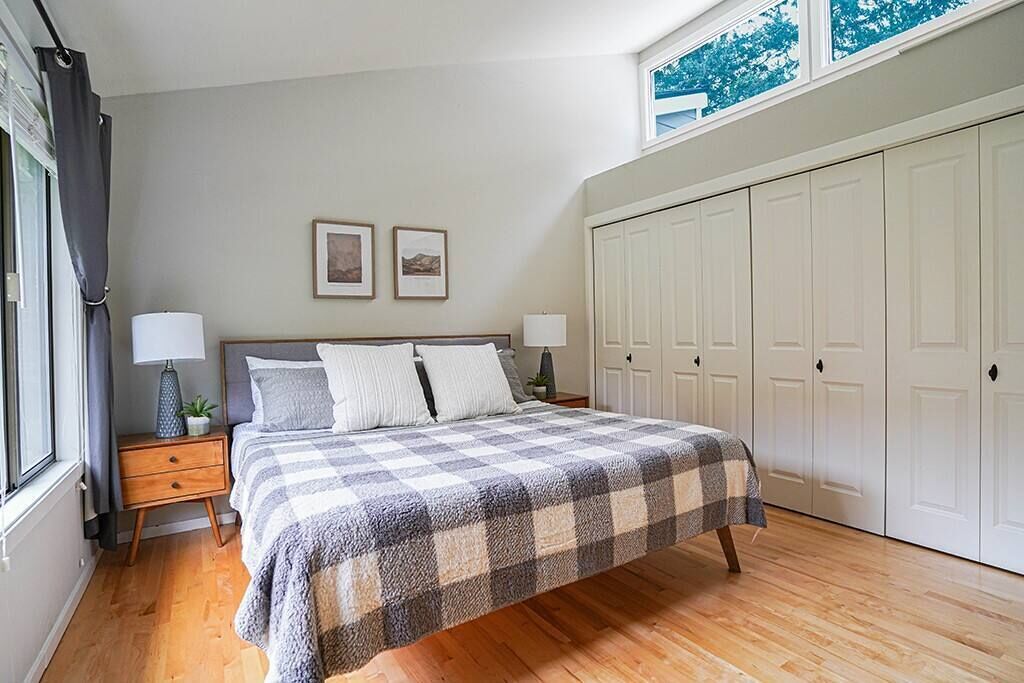 A bedroom with a checkered bedspread, two bedside lamps, a nightstand, a window with curtains, and a wardrobe.