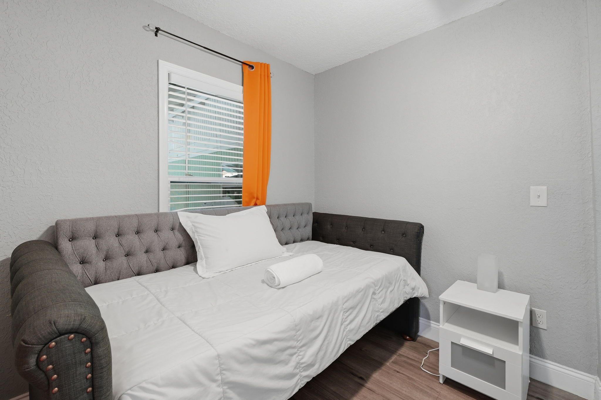 A bedroom with a grey upholstered bed, white bedding, a white bedside table, and a window with orange curtains.