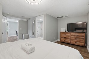 A bedroom with a large bed, a wooden dresser, a flat-screen TV, and a bathroom visible through an open door.