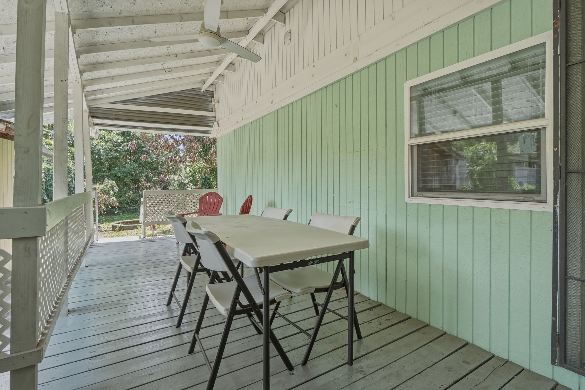 A covered outdoor patio with a table and chairs, a window, and a view of greenery.