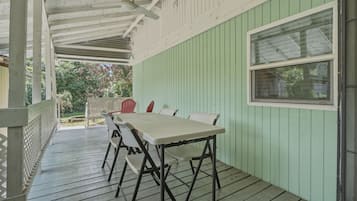A covered outdoor patio with a table and chairs, a window, and a view of greenery.