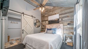 A bedroom with a bed, a ceiling fan, a wooden wall, a window, and a door. - Romantic Waterfall cabin-Hot Tub-Coffee & Tea Bar-Fire Pit (Hot Springs)