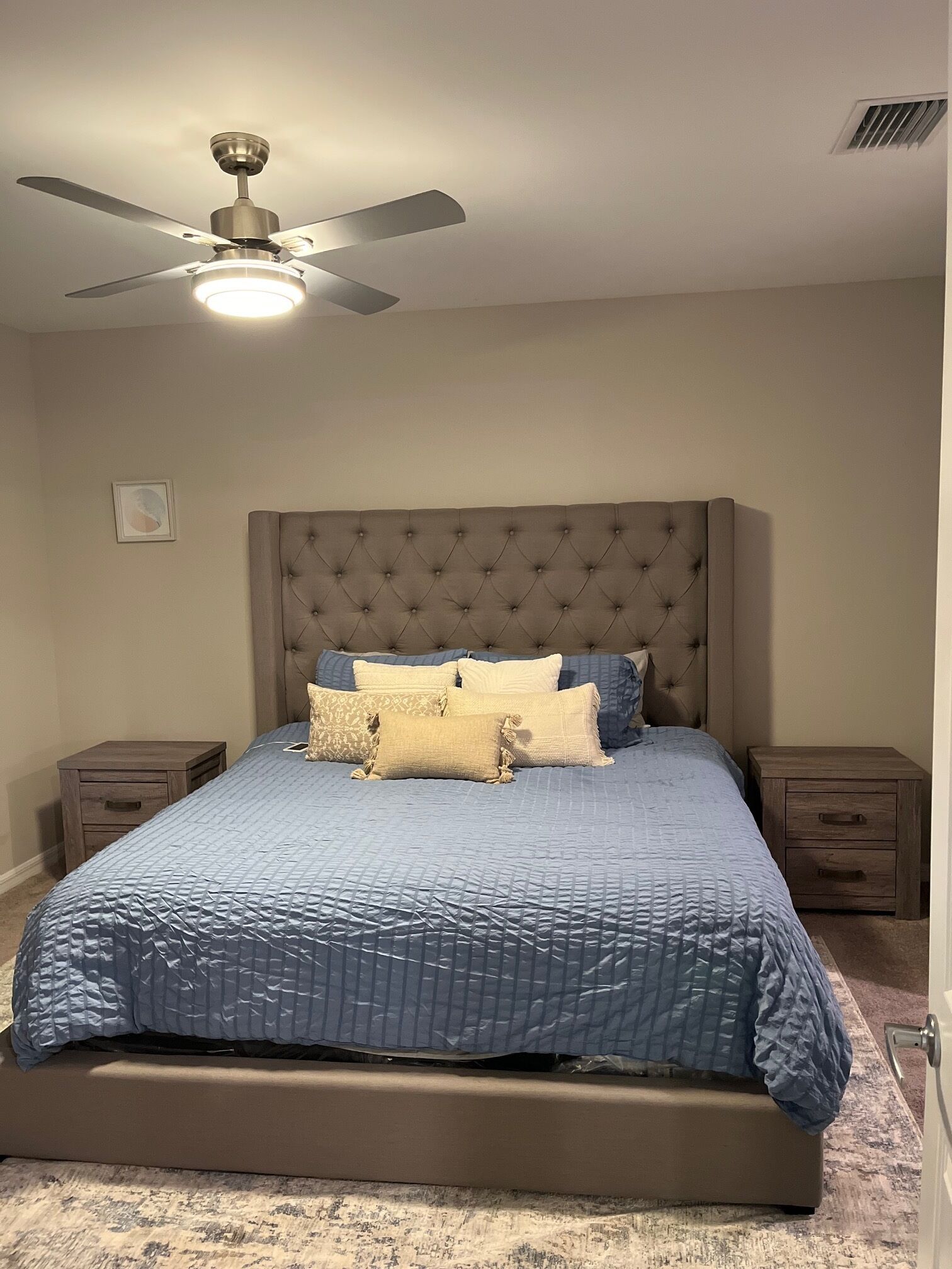 A bedroom with a tufted headboard, a bed with a blue quilt, two bedside tables, and a ceiling fan.