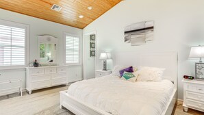 A bedroom with a wooden ceiling, white walls, a large bed with white bedding, a dresser, a nightstand with a lamp, and a mirror. - Waterfront Stuart Oasis w/ Hot Tub & Dock! (Stuart)