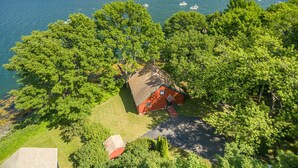 A red cabin with a sloped roof surrounded by lush green trees, overlooking a large body of water with several boats. - Bay-View. Waterfront Log cabin on the bay. Pet Friendly.  (Harpswell)