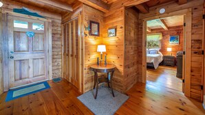 A wooden cabin interior with a door, a small table with a lamp, and a bedroom with a bed and dresser. - Bay-View. Waterfront Log cabin on the bay. Pet Friendly.  (Harpswell)