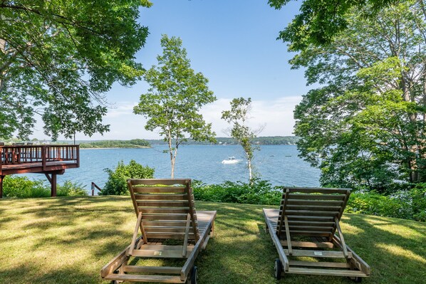 Two wooden lounge chairs facing a lake with a wooden deck and railing in the background. - Bay-View. Waterfront Log cabin on the bay. Pet Friendly. (Harpswell)