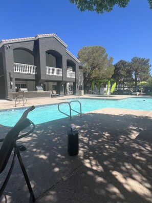 A pool area with a large building in the background, a lounge chair, and a trash can. - Shared apartment wit pool and hot tub (Las Vegas)