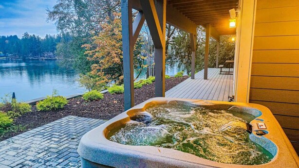 A hot tub with a view of a lake and trees, surrounded by a wooden deck and a covered area. - Modern Shelton Waterfront:Hot Tub,Games&Serenity (Shelton)