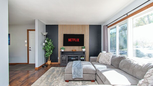 Una sala de estar moderna con un sofá gris, un televisor de pantalla plana en la pared, una planta en maceta y un gran ventanal que proporciona luz natural.