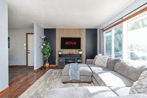 A modern living room with a grey sofa, a flat-screen TV mounted on a wall, a potted plant, and a large window providing natural light. - 4BR Fenced Yard I King Bed, Games, Pack n Play (West Fargo)