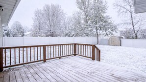 Un patio trasero nevado con una terraza de madera, una cerca blanca y un cobertizo.