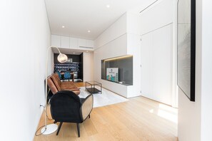 Een moderne woonkamer met een bank, een zwarte fauteuil, een glazen salontafel en een aan de muur bevestigde televisie.