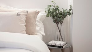 A neatly made bed with white bedding and a beige pillow, a small bedside table with a book and a potted plant. - GuestReady - Mompilher Apartments (Porto)
