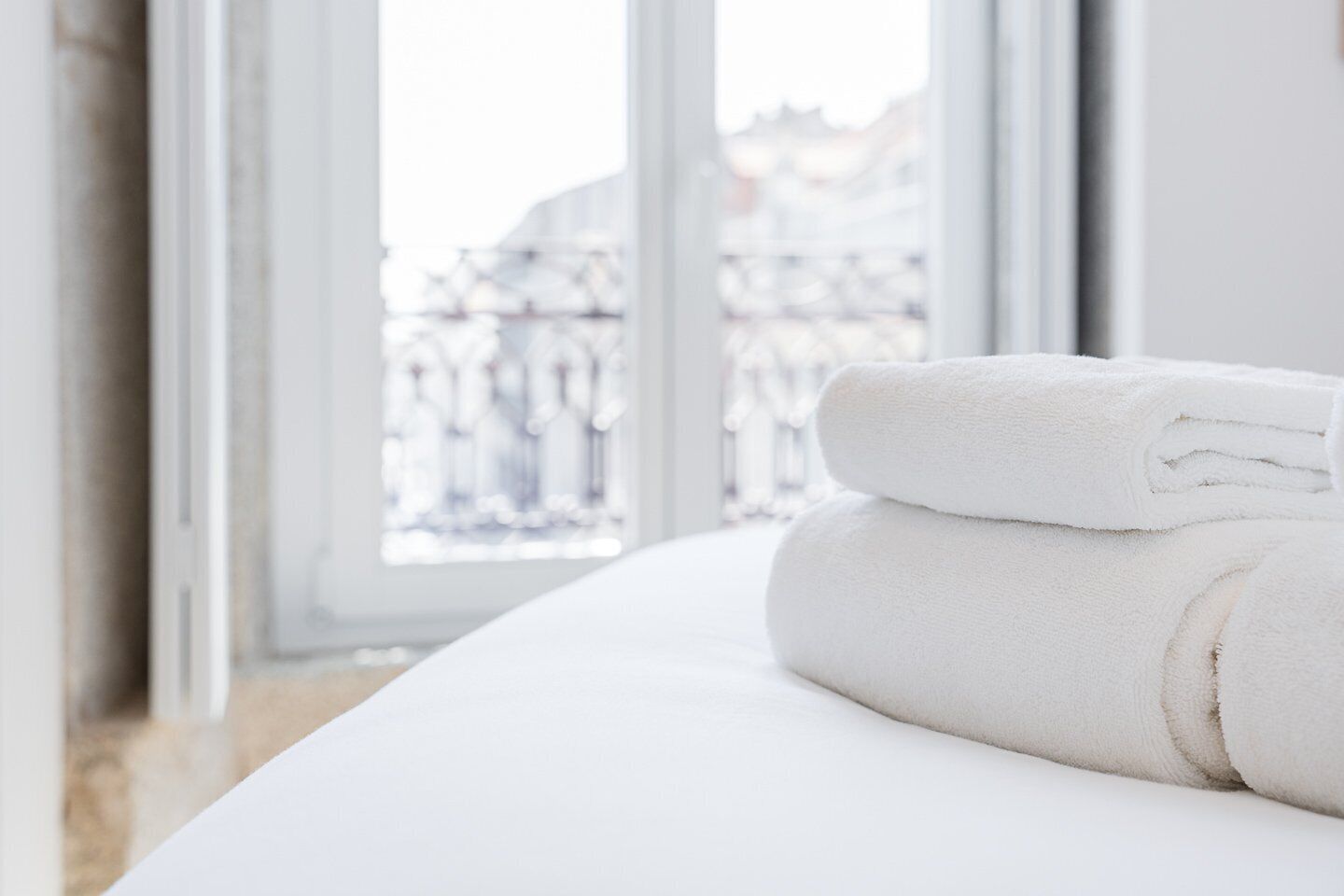 A neatly made bed with white linens and a folded towel on top, set against a window with a view of buildings.