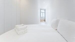 A modern, minimalist bedroom with a neatly made bed, a stack of folded towels, and a window with a view of a balcony. - GuestReady - Mompilher Apartments (Porto)