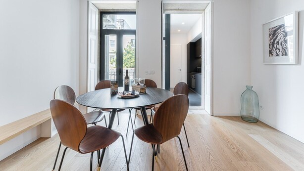 A modern dining room with a round table, six wooden chairs, and a view of the city through large windows. - GuestReady - Mompilher Apartments (Porto)