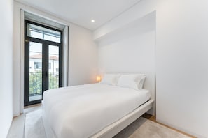 A modern bedroom with a large bed, a bedside lamp, and a view of the outside balcony. - GuestReady - Mompilher Apartments (Porto)