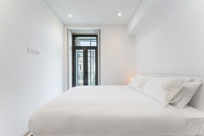 Un dormitorio minimalista con una cama grande, una ventana que ofrece vistas a edificios y una lámpara en el techo.