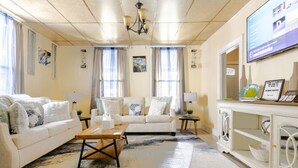 Ein Wohnzimmer mit Sofa, Couchtisch und Fernseher. Vorhänge, eine Chandelierleuchtre und Kunstwerke schmücken die Wände.