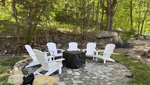 A circular outdoor seating area with white Adirondack chairs and a table covered with a black tablecloth, surrounded by a stone wall and greenery.