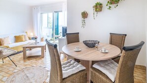 A cozy living room with a round wooden table, wicker chairs, and a sofa. There are hanging plants on the wall and a view of the outdoors through large windows.
