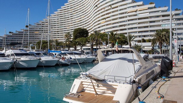 A marina with multiple yachts docked, a large modern building with balconies, and palm trees in the background.