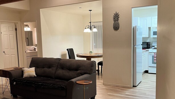 A modern living room with a dark sofa, a small round table, and a patterned rug. There is a dining area with a chandelier and a kitchen visible through an open door.