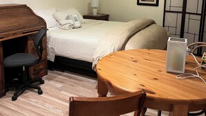 A hotel room with a bed, a desk with a chair, a wooden table with a chair, and a bedside table with a lamp.
