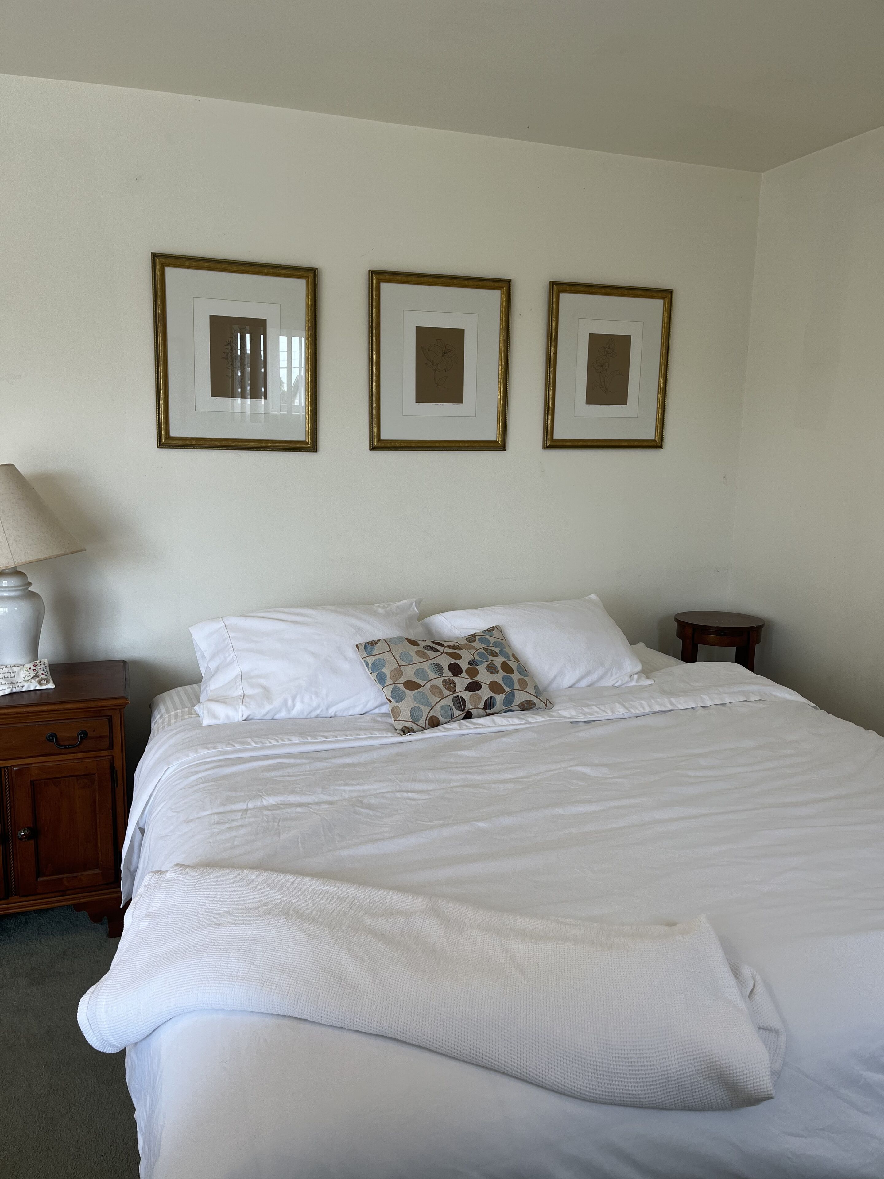 A neatly made bed with white bedding, a patterned pillow, and a lamp on a wooden nightstand. Three framed pictures are hung on the wall above the bed.