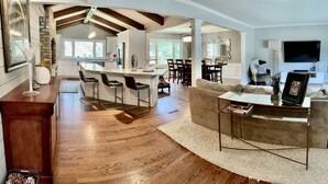 A spacious living area with a large sofa, a coffee table, and a TV. There is a kitchen in the background with a bar and chairs.