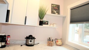 Una cocina con armarios blancos, una cafetera, una cocedora de arroz y una ventana con persianas.