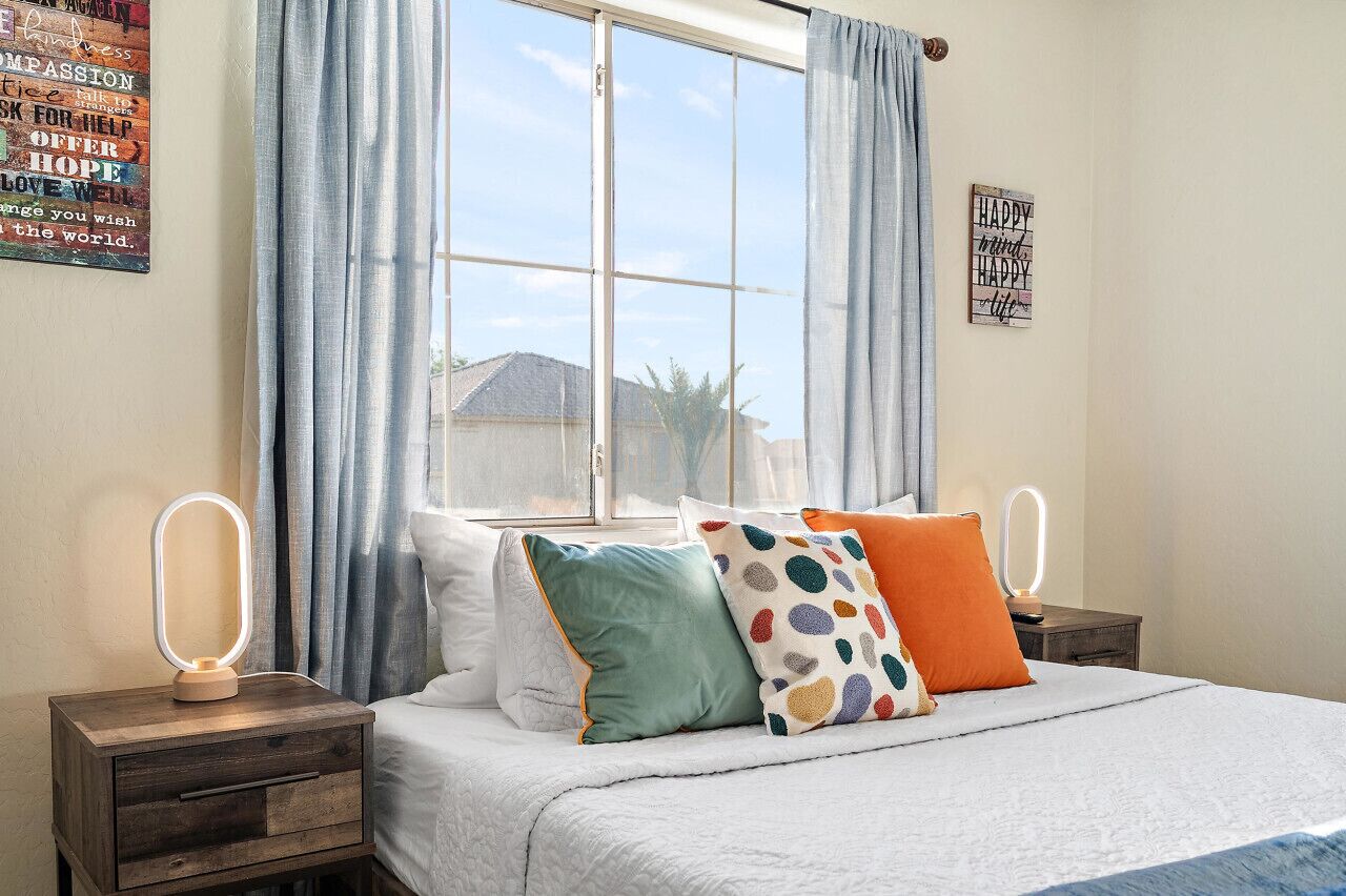 A bedroom with a large window, a bed with multiple pillows, two bedside tables with lighted mirrors, and motivational posters on the walls.