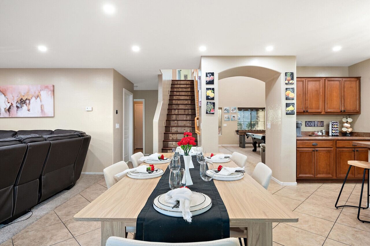 Uma sala de jantar com uma mesa de madeira posta para quatro pessoas, rodeada por cadeiras brancas, e um jogo americano preto. Uma cadeira de massagem é visível ao fundo.