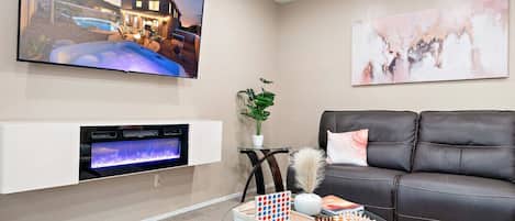 A living room with a fireplace, a black leather sofa, a glass coffee table with a chessboard, and a TV mounted on the wall displaying a sunset scene.