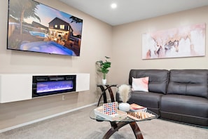 A living room with a fireplace, a black leather sofa, a glass coffee table with a chessboard, and a TV mounted on the wall displaying a sunset scene.