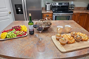A kitchen counter with a fruit platter, wine glasses, a bottle of wine, and muffins.