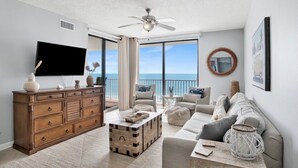 Een ruime woonkamer met een grote flatscreen-tv, een houten dressoir, een zithoek en uitzicht op de oceaan door grote ramen.