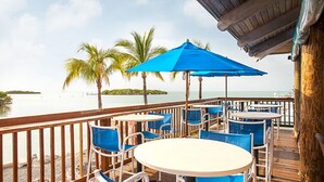 A deck with tables and chairs, overlooking a calm body of water with palm trees.