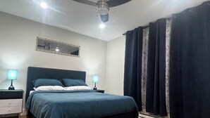 A bedroom with a bed, two bedside tables, a ceiling fan, a mirror, and dark curtains. - City Center Modern Apartment (Charlottetown)