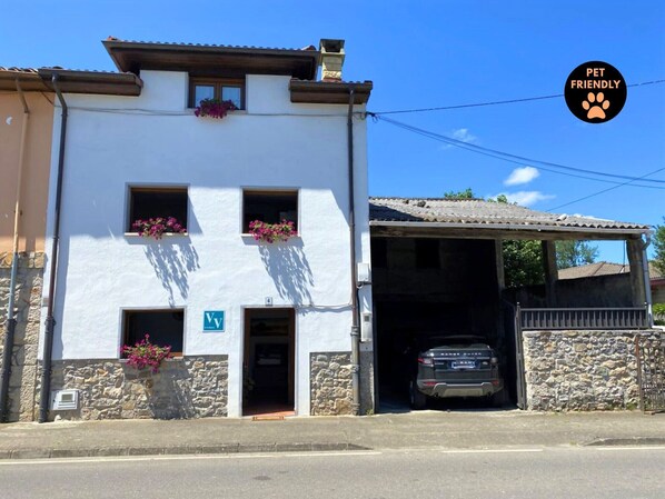 A building with a white facade, two windows with flower boxes, a visible 'V' sign, and a parked car in the background.