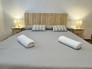 Cama bem arrumada com colcha cinza texturizada, dois travesseiros e duas toalhas enroladas. Há duas mesas de cabeceira com abajures e uma cabeceira de madeira.