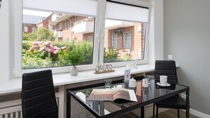 A modern dining area with a glass table, black chairs, and a view of a garden with pink flowers through large windows. - Adrian 3 - Gemütliches, Zentrales Apartment für 2 Personen, Wlan und TV, Separate Küche (Sylt)